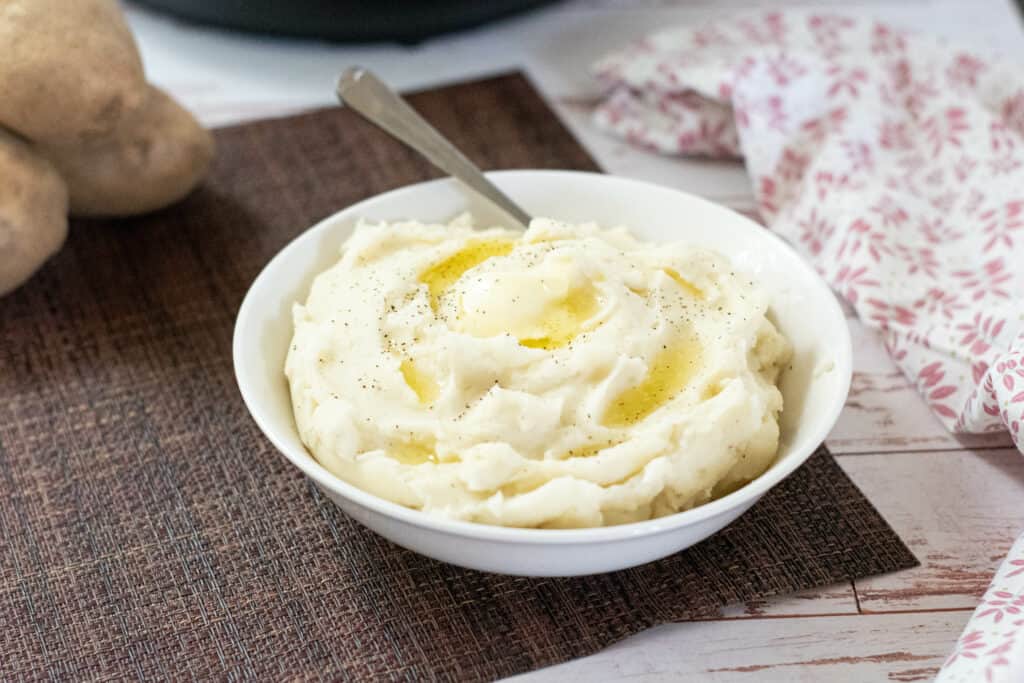 White bowl of mashed potatoes with a butter on top and a spoon in the bowl.  There are potatoes and a linen in the background 