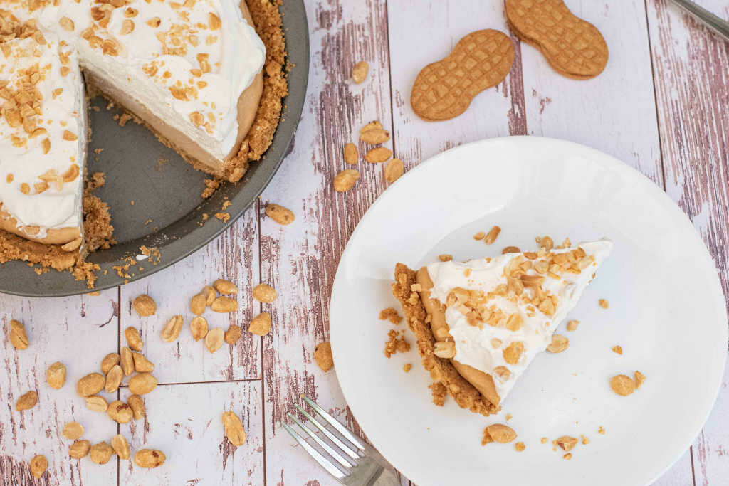 Piece of peanut butter pie on a white plate and pie with a piece missing surrounded by peanuts and Nutter Butter cookies 