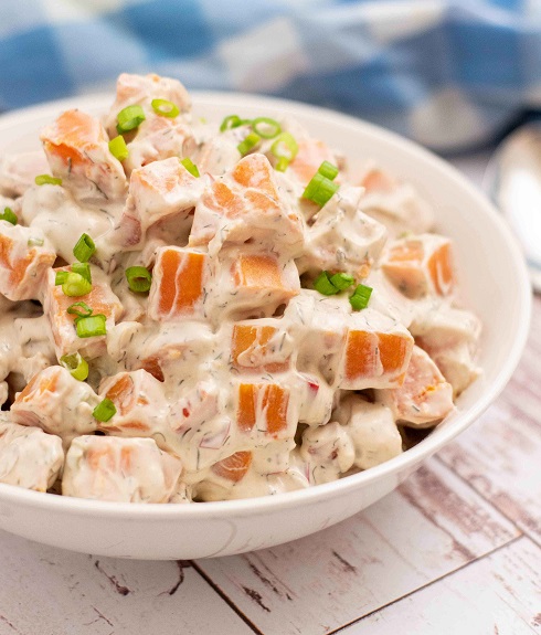 sweet potato salad in a white bowl garnished with green onions on a blue and white checkered table cloth 