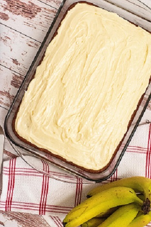 An entire banana cake with cream cheese frosting in a glass rectangle pan. There is a bunch of bananas in the corner and a red and white linen