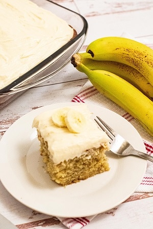 A slice of banana cake on a white plate with a bunch of bananas beside it and a cake pan on the other side 