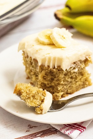 A side view of a piece of banana cake on a white plate with a fork with a bite of cake on it. The cake has sliced banana on top 