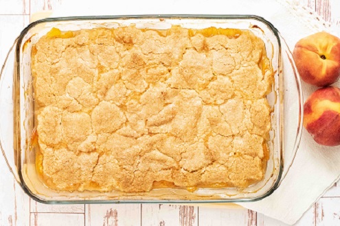 Baked peach cobbler in a glass pan on a white cloth with peaches on the side