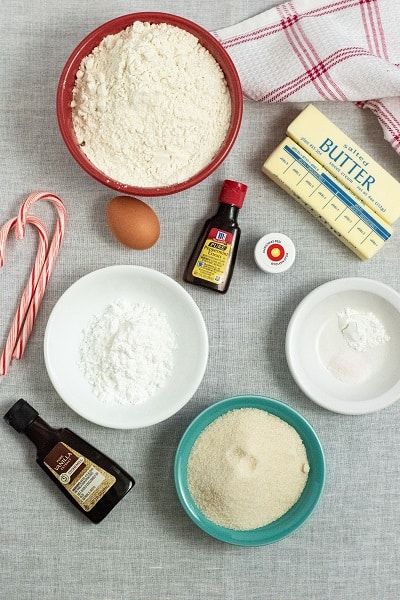 Ingredients for candy cane cookies, flour, egg, butter, peppermint extract, vanilla extract. baking powder, sugar, powdered sugar, red food coloring and candy canes all laid out on a white background 