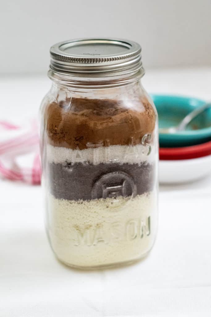 A mason jar with ingredients layered in it. There is a pile of empty bowls and a red and white linen in the background. 