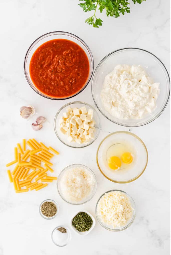 Clear bowls with the ingredients for baked ziti on a white background, Ricotta cheese, mozzarella, eggs, garlic, parmasean, italian seasoning, parsley, salt, pepper, and dried ziti