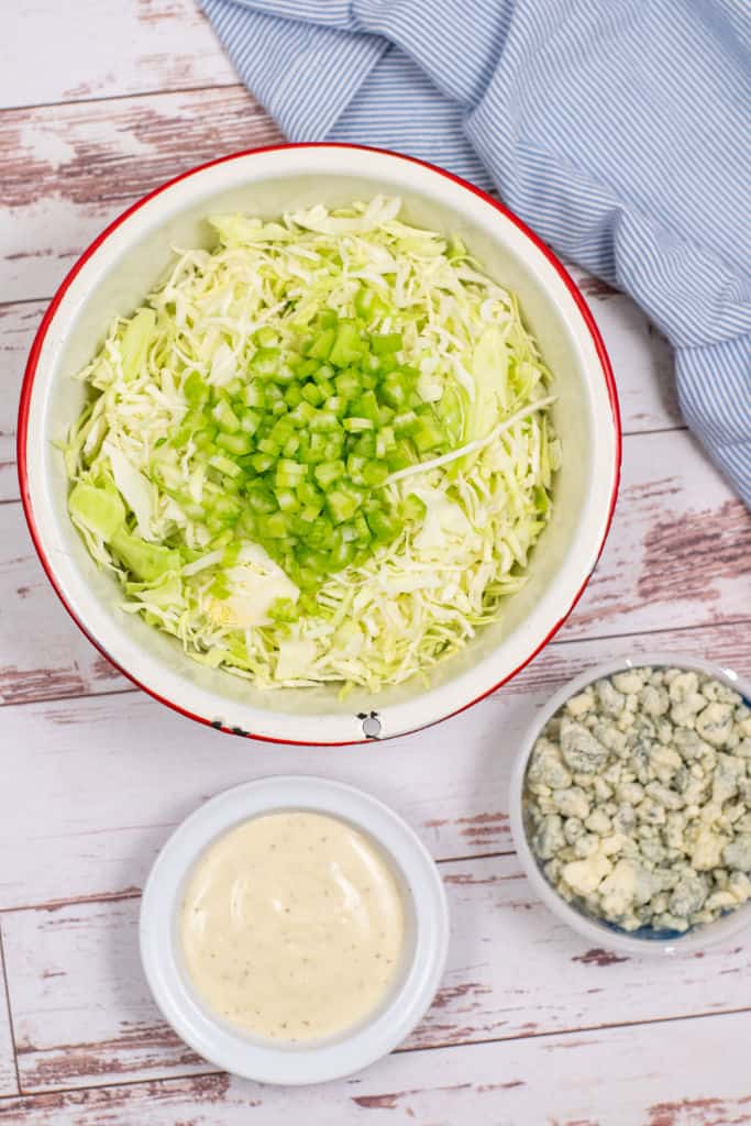 A white bowl with shredded cabbage and diced celery in it. 2 smaller bowls, one with blue cheese dressing and one with crumbled blue cheese. 