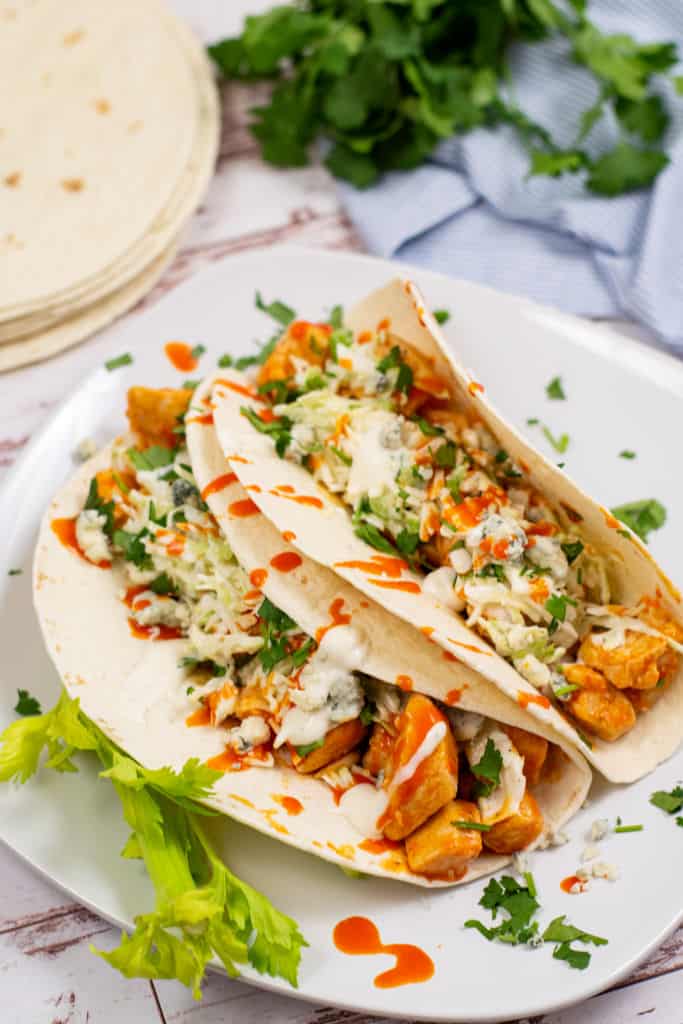 Two tacos on a white plate. Flour tortillas filled with buffalo chicken, coleslaw, blue cheese, and buffalo sauce. A stack of tortillas and a bunch of cilantro are in the background. 