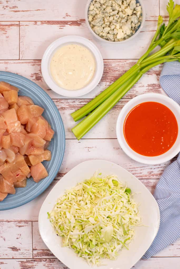 Ingredients for buffalo chicken tacos, cubed chicken breast, blue cheese dressing, blue cheese, buffalo wing sauce, coleslaw mix and 2 stalks of celery on a white wooden background. 