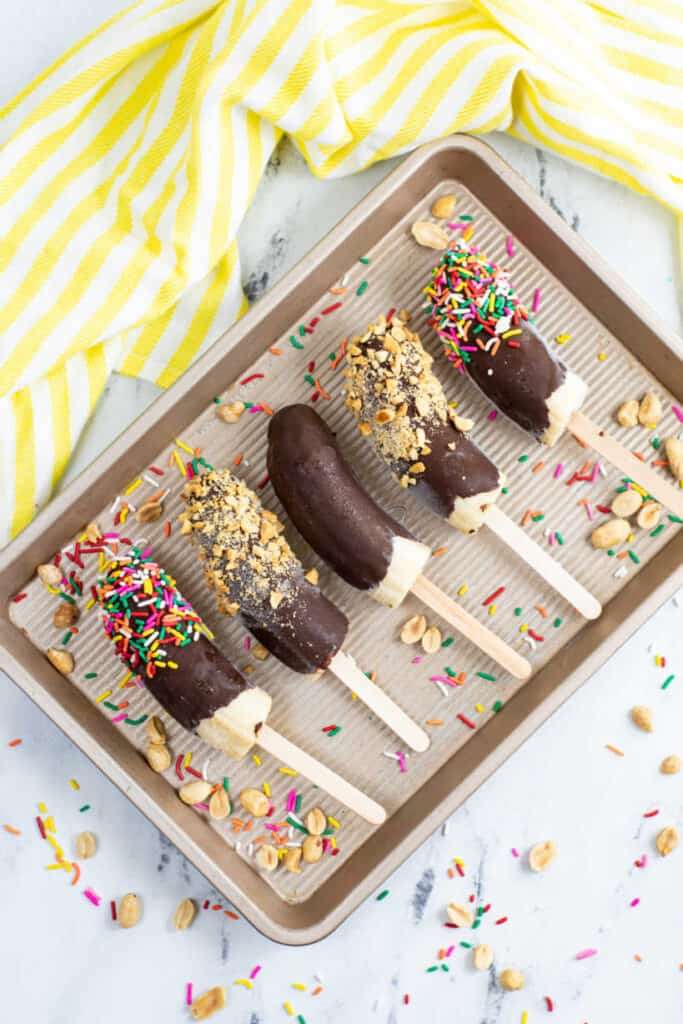 5 chocolate covered bananas with chopped nuts and sprinkles on a small metal tray 