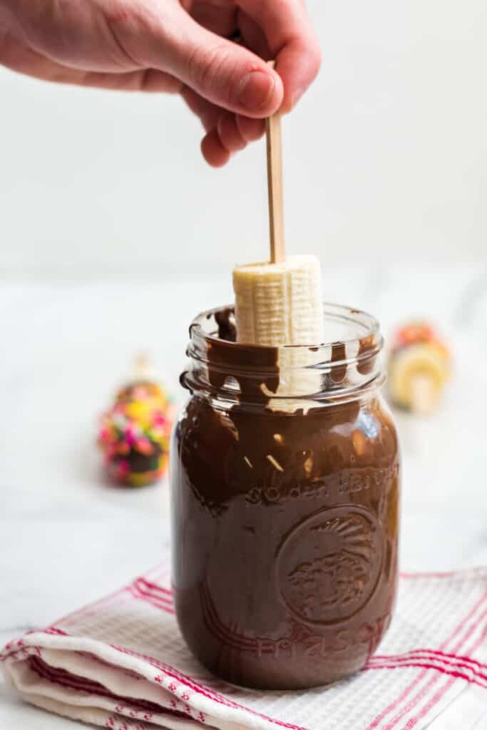 A hand dipping half of a banana on a popsicle stick into a Mason jar full of melted chocolate 