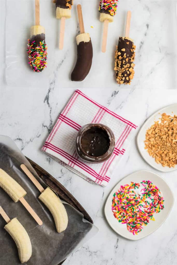 a counter top with a tray full of frozen banana halves on popsicle sticks, melted chocolate, plates with sprinkles and nuts, and a piece of wax paper with finished banana pops on it. 