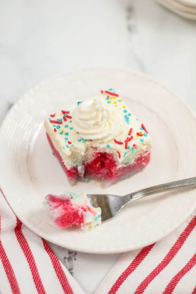A piece poke cake with white frosting an sprinkles on a white plate with a fork taken out. 
