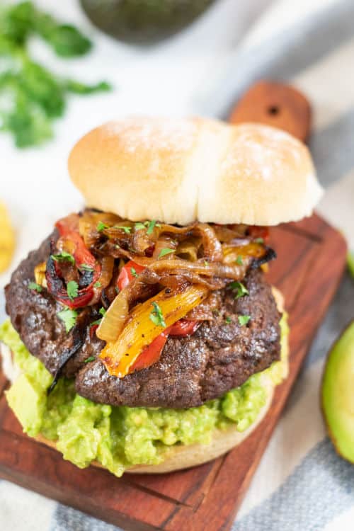 A burger on a bun with guacamole topped with peppers and onions