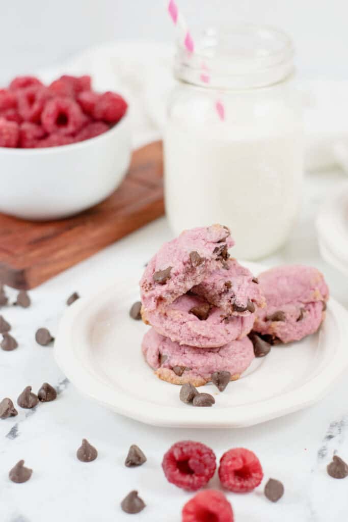 a white plate with a stack of cookies on it, with a mason jar full of milk in the background.  There are chocolate chips and raspberries scattered around. 