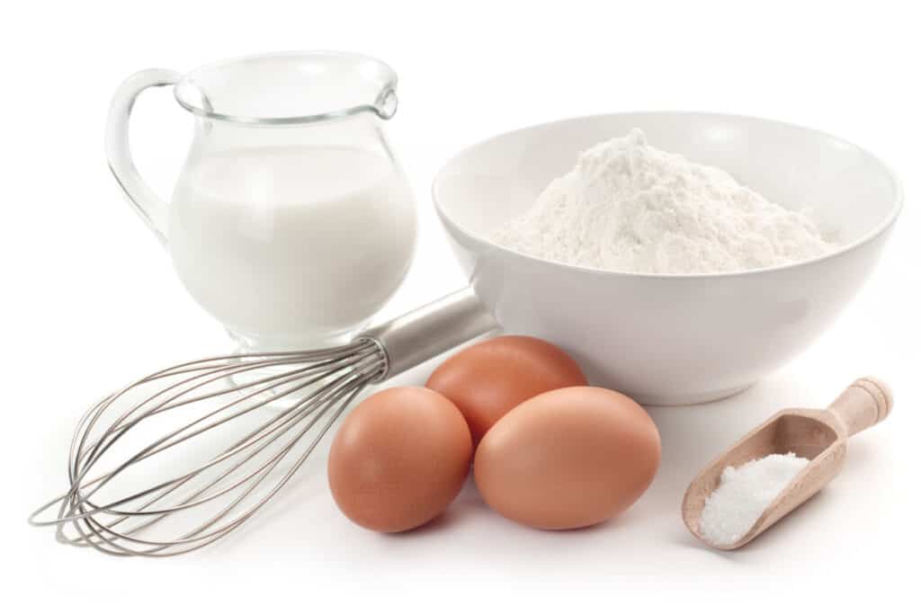 A pitcher of milk, bowl of flour, whisk 3 brown eggs and scoop of salt 