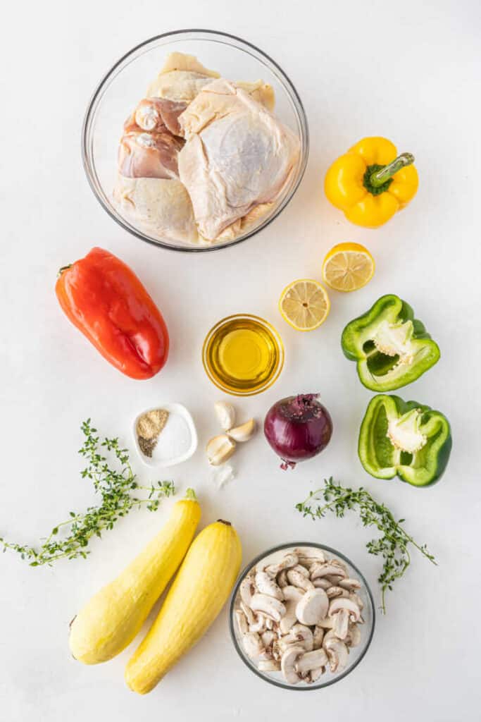 The ingredients for chicken and vegetables sheet pan dinner laid out on a white background. Chicken thighs, mushrooms, green, red and yellow peppers, red onion, lemon, yellow squash, fresh thyme, garlic cloves, and spices. 