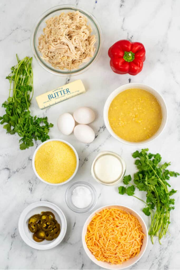 The ingredients for cornbread casserole on a white marble background. Shredded chicken, red pepper, butter, eggs, cilantro, cream corn, corn meal, milk, pickled jalapenos and shredded cheddar cheese 