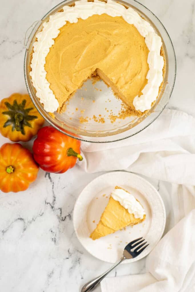 A pumpkin pie with a piece removed, next to it is a white plate with a piece of pie on it. 