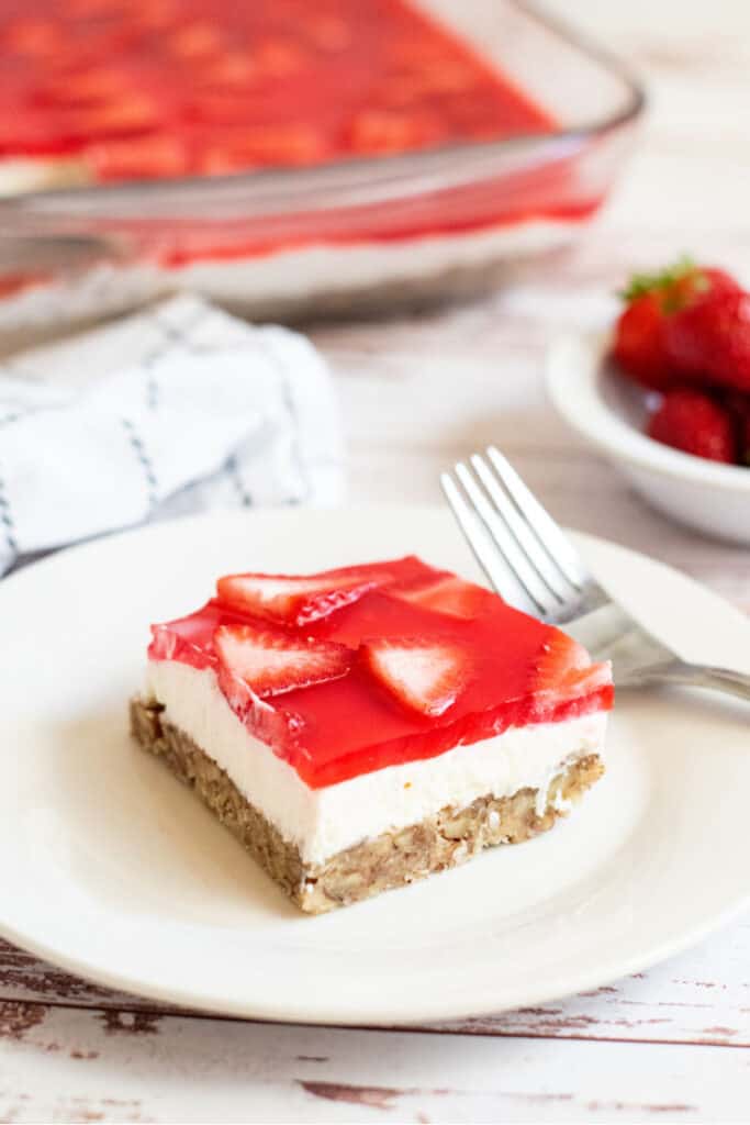 A white plate with a piece of keto strawberry pretzel salad, there is a dish of strawberries on the background. 
