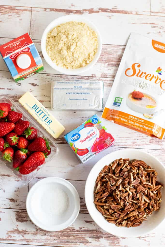The ingredients for keto strawberry pretzel salad on a weathered white wood surface.  Heavy whipping cream, almond flour, cream cheese, Swerve sweetener, butter, sugar free strawberry jello, strawberries, pecans and salt. 