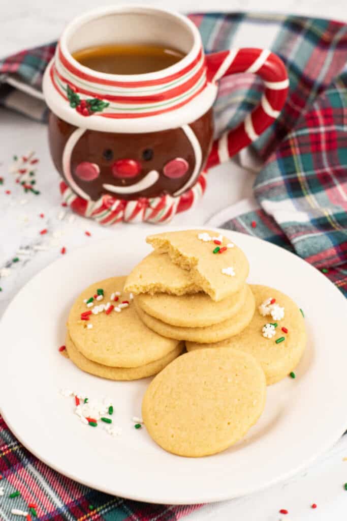 a pile of sugar cookies on a white plate, there is gingerbread man mug in the background. 