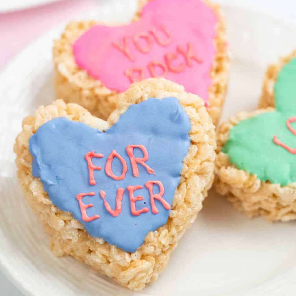A plate full of conversation heart rice krispie treats. There is one in focus that is blue and says "For Ever" 