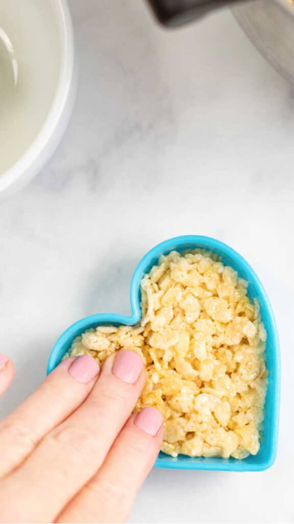 A hand pressing rice krispie treat mixture into a heart shaped cookie cutter. 