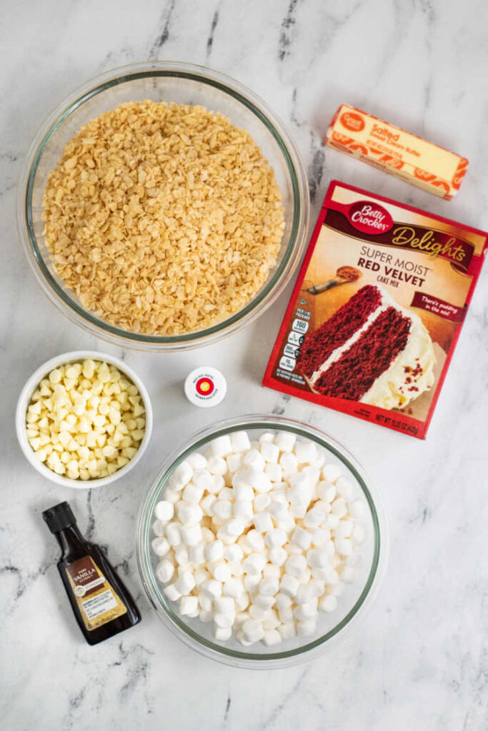 The ingredients for red velvet rice krispie treats on a marble countertop. Crispy rice cereal, red velvet cake mix, marshmallows, butter, vanilla, white chocolate chips, gel food coloring. 