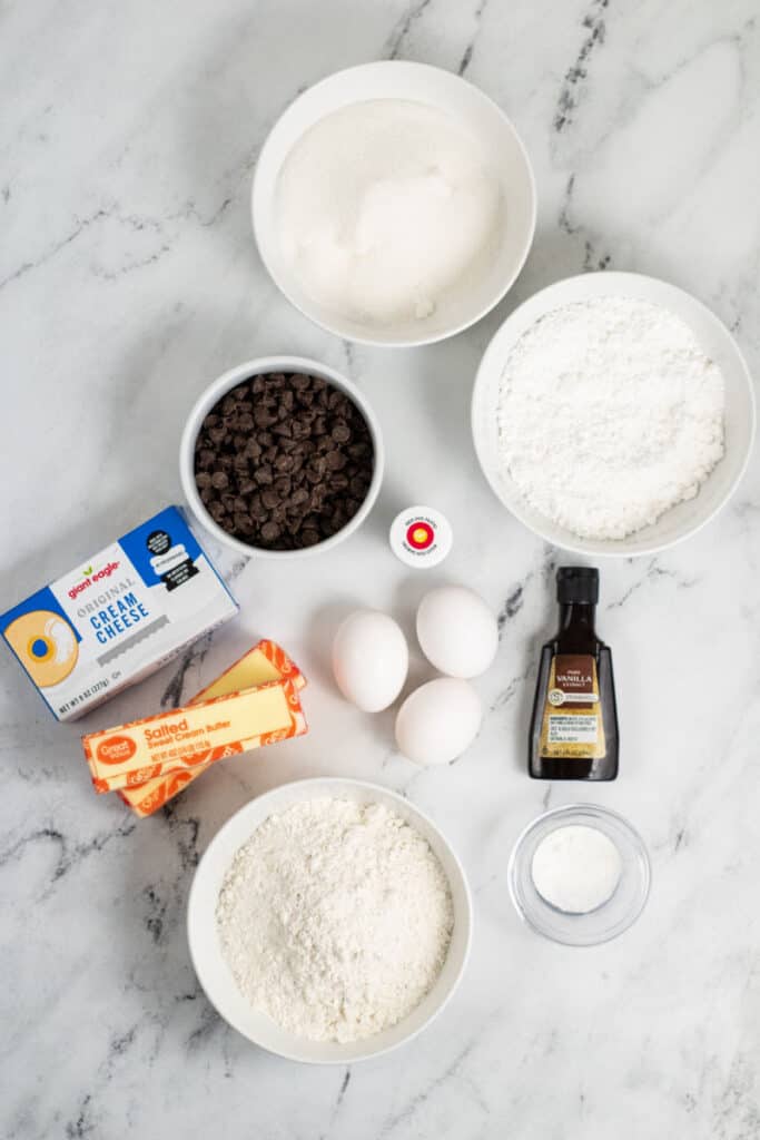 The ingredients for red velvet brownies with cream cheese frosting. A bowl of sugar, flour, chocolate chips, red food coloring, vanilla, cream cheese, butter, powdered sugar, baking powder and salt 