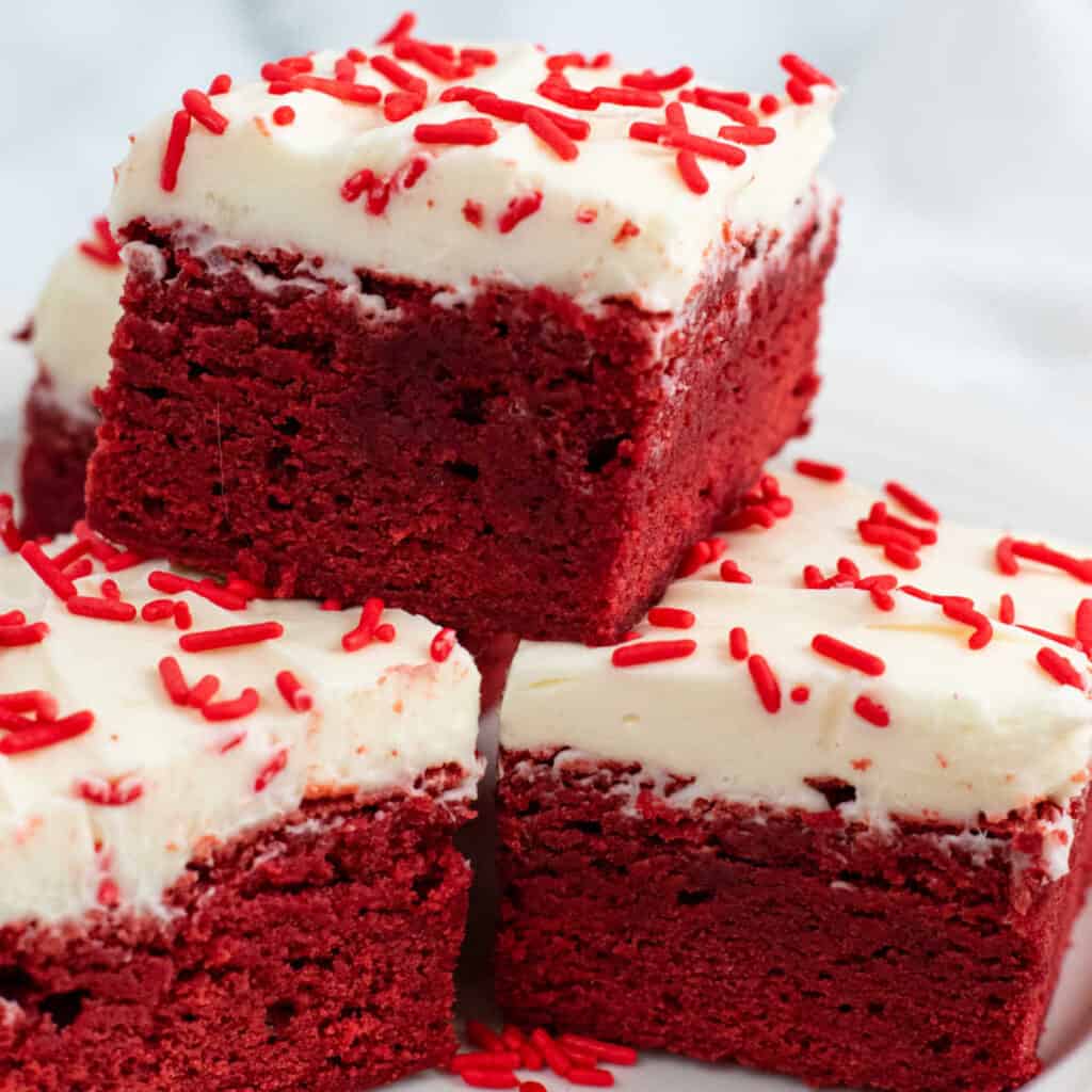 A stack of red velvet brownies frosted with cream cheese frosting and sprinkled with red sprinkles 