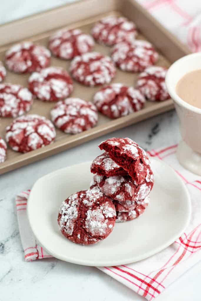 A stack of red velvet cookies on a white plate. Behind them is a baking sheet with more cookies. 