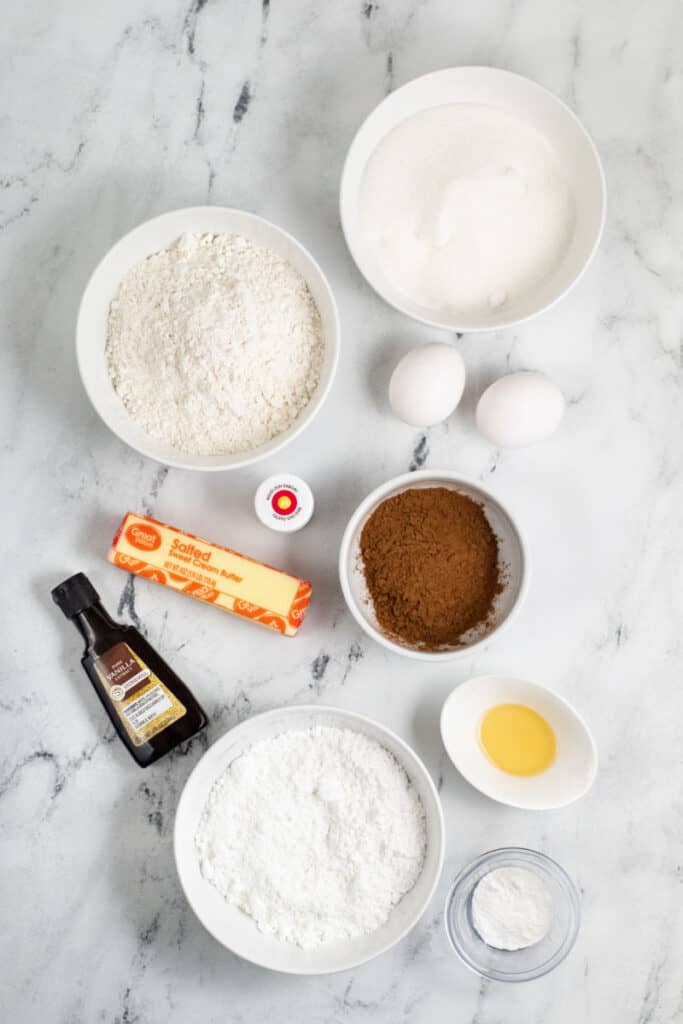 The ingredients for red velvet crinkle cookies laid out on a marble counter top. There is flour, eggs, butter, cocoa powder, vanilla extract, powdered sugar, baking soda and apple cider vinegar and red food coloring 