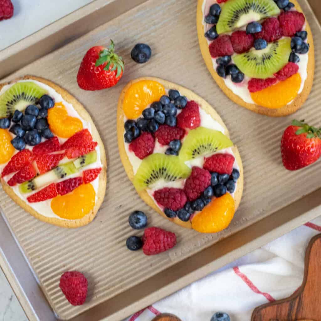 Egg shaped sugar cookies frosted with white frosting and decorated with fruit on a metal baking sheet. 