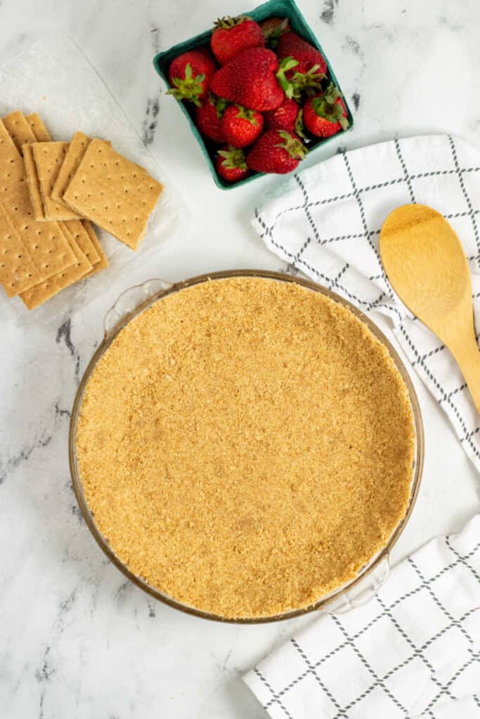A graham cracker crust in a glass pie pan, next to it is a container of strawberries and a wooden spoon. 