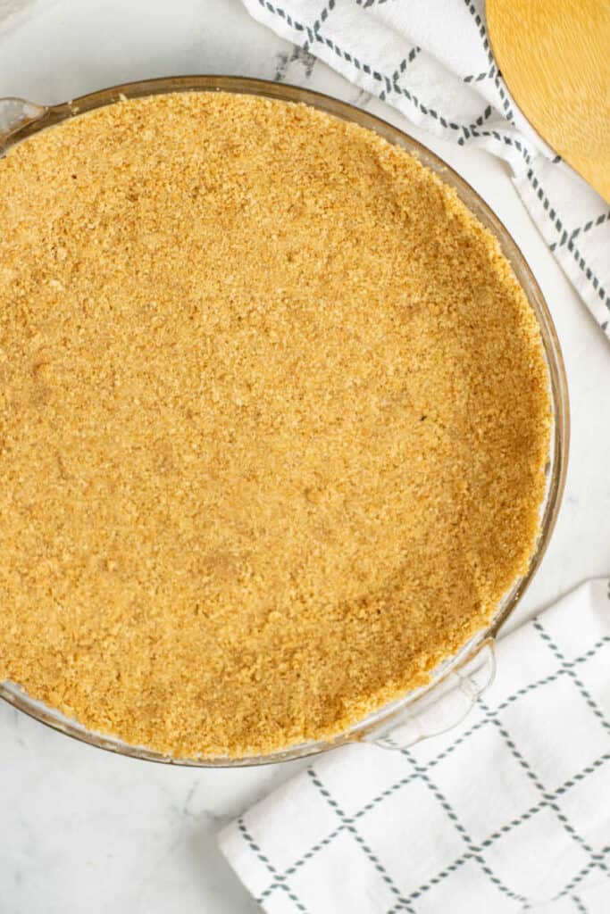 A glass pie pan with a graham cracker crust on a white marble countertop. 