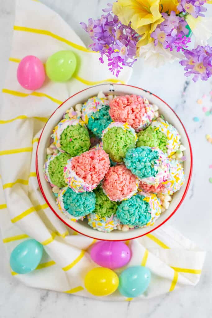 A white bowl full of Easter egg rice krispie treats. It is surrounded by colorful plastic Easter eggs on a white marble counter top. 