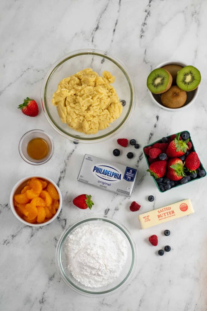 The ingredients for fruit pizza cookies on a marble background. There is sugar cookie dough, cream cheese, butter, vanilla, powdered sugar, berries, kiwis, and mandarin oranges. 