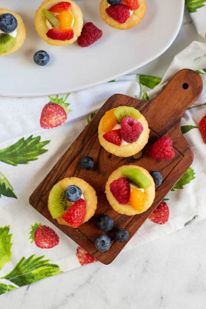 A wooden platter with mini fruit tarts on a linen covered with strawberries. 