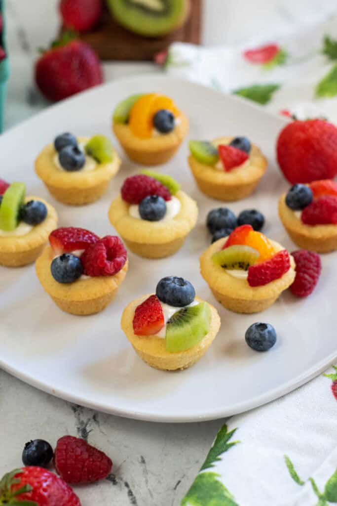 Mini fruit tarts on a white square plate surrounded by berries. 