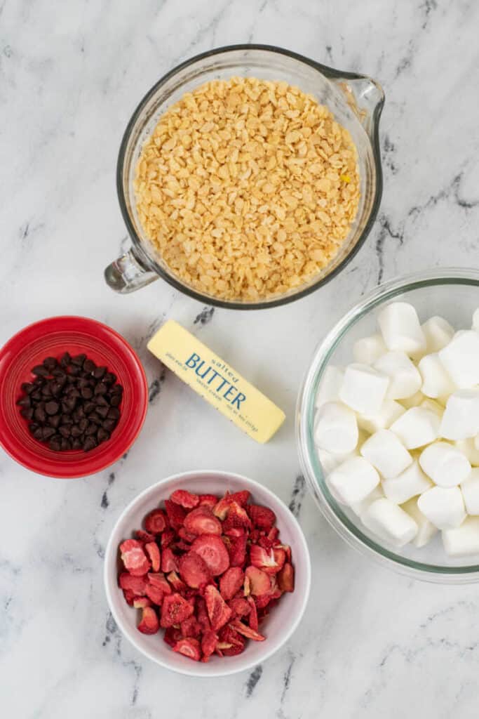 The ingredients for Neapolitan Rice Krispies Treats on a marble counter top. A bowl of rice cereal, a bowl of marshmallows, a stick of butter, chocolate chips and freeze dried strawberries. 