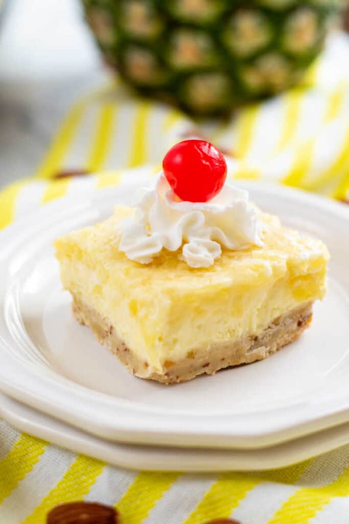 A piece of pineapple cheesecake bar on a white plate topped with whipped cream and a cherry on top. 