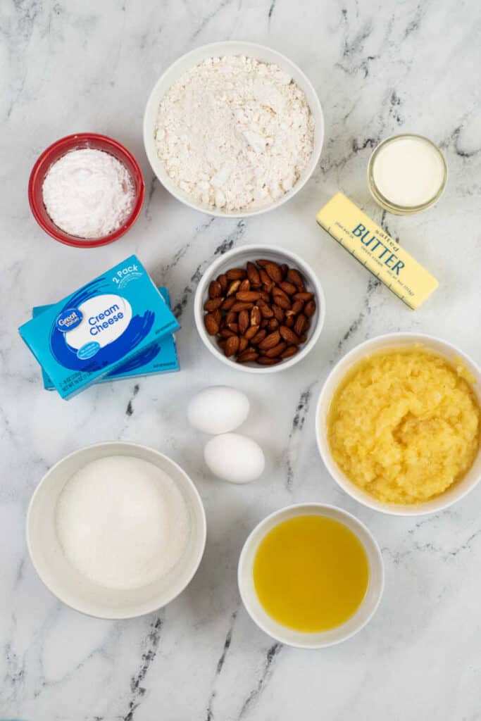 The ingredients for pineapple cheesecake bars on a marble counter top. Flour, powdered sugar, milk, butter,  almonds, cream cheese, eggs, crushed pineapple, sugar and pineapple juice. 