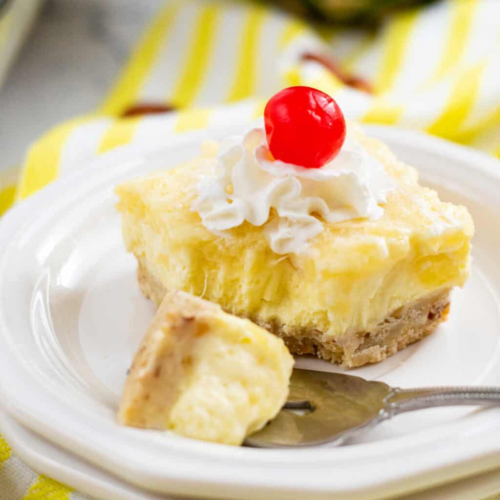 A pineapple cheesecake square on a white plate with whipped cream and a cherry on top. 