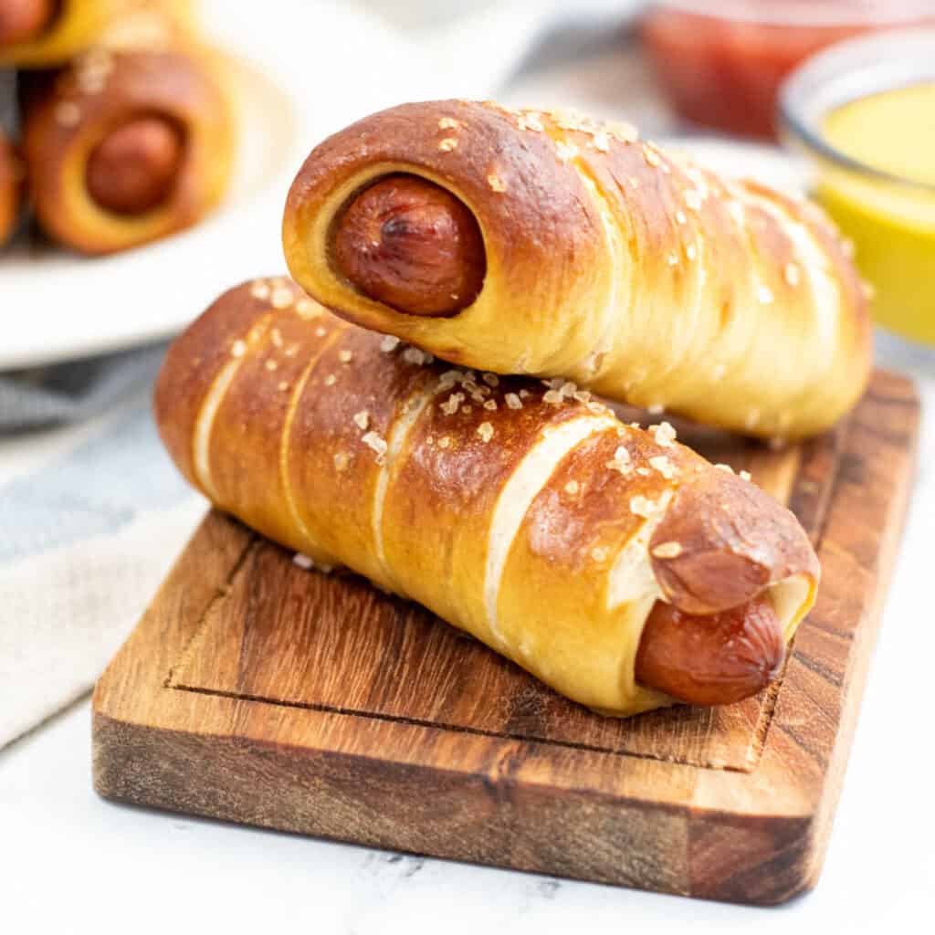Two pretzel dogs on a small wooden chopping block. 