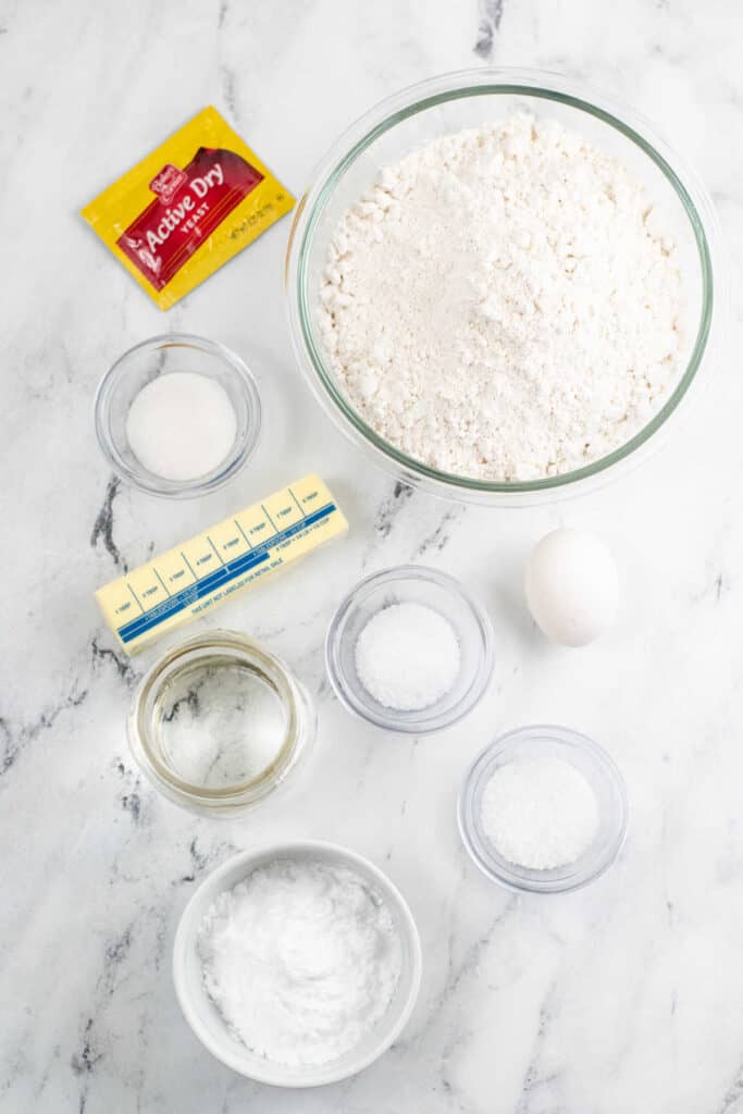 The ingredients to make soft pretzel bites on a marble counter top. Yeast, flour, salt, egg, butter, water, sugar, baking soda, and coarse salt. 