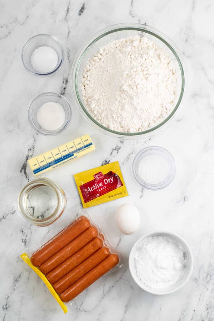 The ingredients for making pretzel dogs on a white marble counter top. Flour, salt, coarse salt, butter, water, egg, yeast, hot dogs, baking soda. 