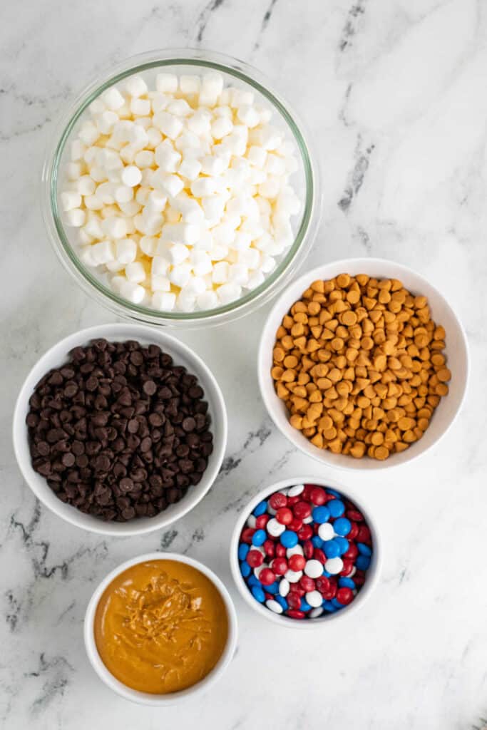 The ingredients for red white and blue marshmallow bars on a white marble counter top. Marshmallows, chocolate chips, butterscotch chips, peanut butter and M&M's 