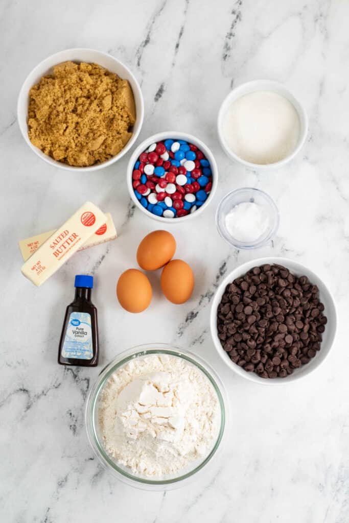 A white marble counter top with the ingredients for cookie bars laid out on it. Brown sugar, white sugar, M&M's baking powder, baking soda and salt, butter, eggs, vanilla, chocolate chips and flour