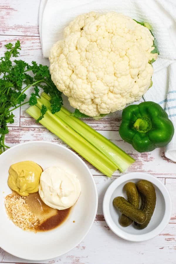 The ingredients for keto cauliflower potato salad. A head of cauliflower, a green pepper, celery, pickles, parsley, mayonnaise, mustard, and spices. 